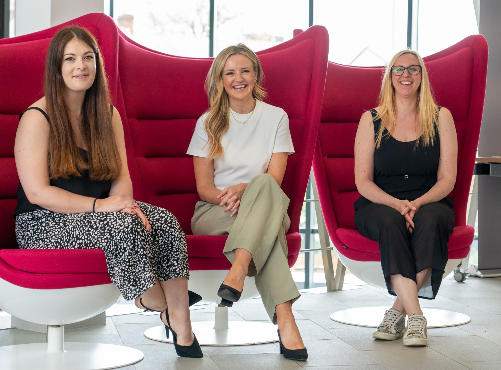 Three women are sitting on big red chairs smiling at the camera