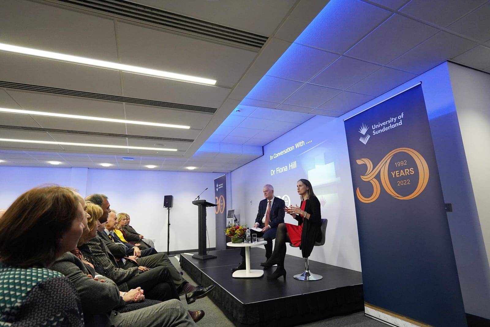 A women in formal red dress and black blazer is giving her speech while everyone below is sitting and listening to her